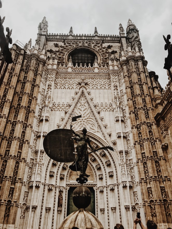The very gothic-looking entrance to the Catedral de Sevilla. There is a statue in front of an angel , who holds a bushel of wheat, and a flag with a banner and what looks like a shield. The catedral itself is huge - it's nearly 16 stories tall!