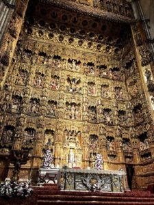 Gold, textured wall and beautifully ornamental main altar in the cathedral. There is a table at the altar with flowers and figurines. In the gold and grand wall behind it are carved images and figurines or major saints and people in the Catholic religion.