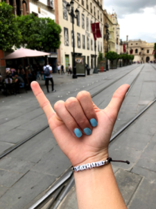 Picture of one of the student's hands, which is making a "Hang-ten" sign, with their thumb and pinky fingers extended, and the rest pulled in to their palm. On the student's wrist is a braclet, which has white block beads which spell out "Sevilla"