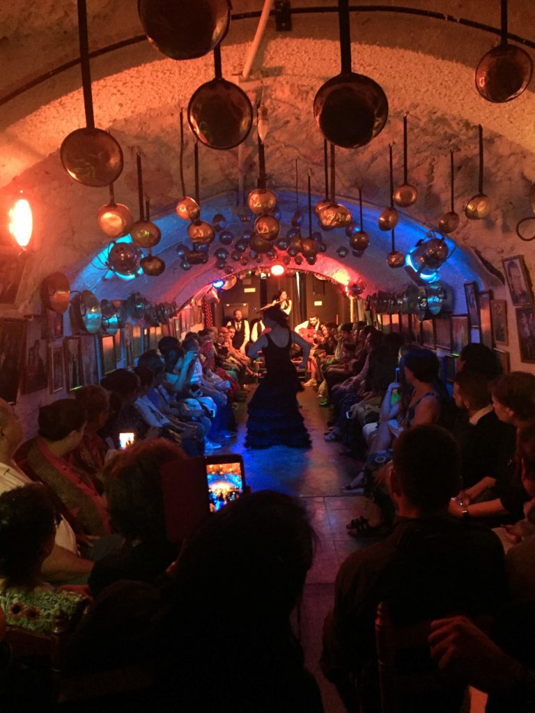 A long, skinny, low roofed room with copper pans hanging from the ceiling. Along the walls sit all the audience members, and in the middle aisle is a female flamenco dancer in a long black dress striking a pose.